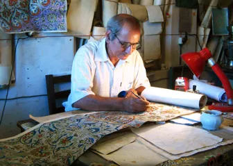 Man designing a rug in his studio. Behind him many scrolls with many different designs can be seen